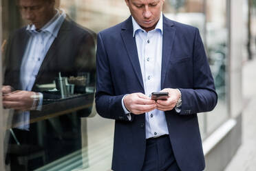 Nachdenklicher reifer Geschäftsmann, der ein Smartphone benutzt, während er vor einem Glasfenster steht - MASF16927