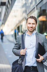 Portrait of smiling mature entrepreneur with bag standing outdoors - MASF16958