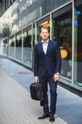 Porträt eines reifen Geschäftsmannes mit Tasche, der vor einem Bürogebäude steht - MASF16965