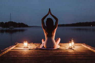 Rückansicht einer Frau, die Yoga praktiziert, während sie auf einem Pier am Meer sitzt, gegen den Himmel - MASF16979