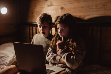 Geschwisterkind benutzt Laptop, während es auf dem Bett an der Wand im Schlafzimmer sitzt - MASF16997