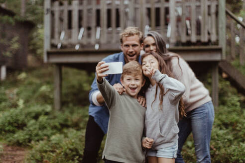 Lächelnde Familie, die ein Selfie mit einem Mobiltelefon macht, während sie vor einem Haus steht - MASF17003