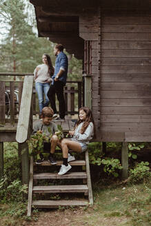 Geschwisterkind in voller Länge auf den Stufen sitzend, während die Eltern in der Veranda stehen - MASF17005