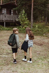 Full length of brother and sister with bag standing against house in forest - MASF17014