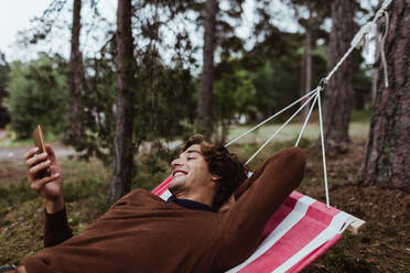 Smiling young man using mobile phone while relaxing in hammock against trees - MASF17028