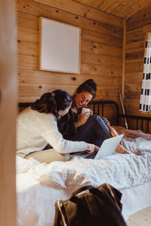 Young woman pointing at laptop while sitting with friend on bed at home - MASF17030