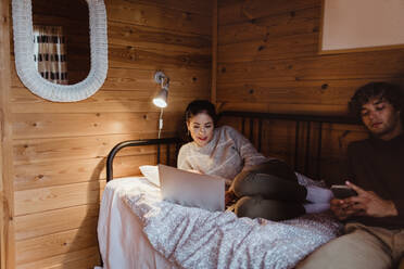 Junge Frau benutzt einen Laptop, während sie mit einem männlichen Freund zu Hause auf dem Bett sitzt - MASF17032