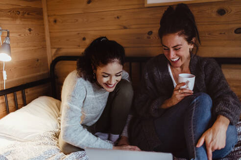 Lächelnde junge Frau, die einen Laptop benutzt, während sie mit einem Freund zu Hause auf dem Bett sitzt - MASF17033