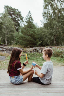 Geschwister essen süßes Essen, während sie auf dem Holzboden vor einem Baum und dem Himmel sitzen - MASF17060