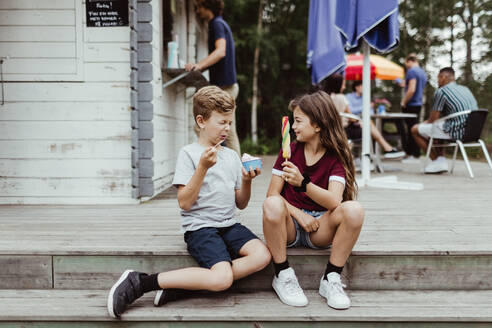 Bruder und Schwester in voller Länge beim Essen von Süßigkeiten, während sie auf Holzstufen sitzen - MASF17061