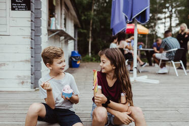 Smiling brother and sister eating sweet food while sitting on wooden floor - MASF17062