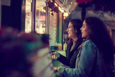 Smiling lesbian couple standing against concession stand in city at night - MASF17113