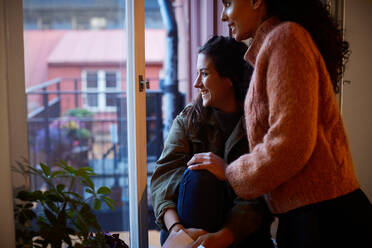 Smiling lesbian couple looking through window at apartment during vacation - MASF17123