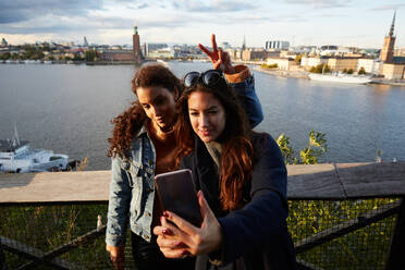 Lesbisches Paar, das ein Selfie mit dem Mobiltelefon an einem Geländer vor einer Stadtlandschaft macht - MASF17132