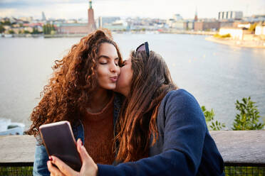 Lesbisches Paar, das sich küsst, während es ein Selfie mit einem Mobiltelefon vor einer Stadtlandschaft macht - MASF17135