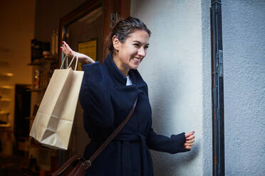 Smiling woman with shopping bag standing against wall in city during vacation - MASF17161