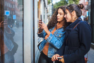 Female friends photographing store window with smart phone in city - MASF17167