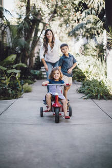 Daughter riding tricycle while mother and brother standing in background - MASF17177