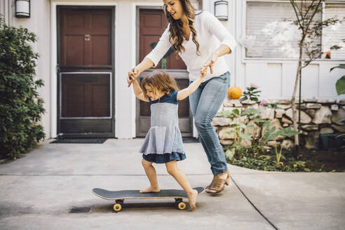 Smiling daughter balancing over skateboard with help of mother on footpath - MASF17181