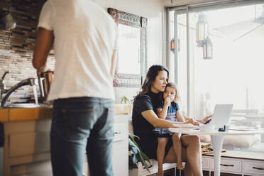 Mother working on laptop while daughter looking at father standing in kitchen - MASF17196