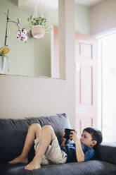 Boy using phone while lying on sofa at home - MASF17198