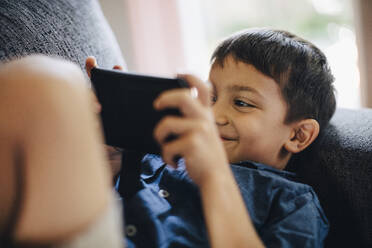 Smiling boy using phone while lying on sofa - MASF17199