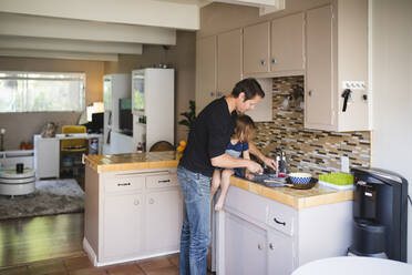 Father washing kitchen utensil while daughter sitting on kitchen counter - MASF17224