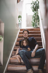 Porträt eines lächelnden Vaters mit Tochter, die zu Hause auf den Stufen sitzt - MASF17230