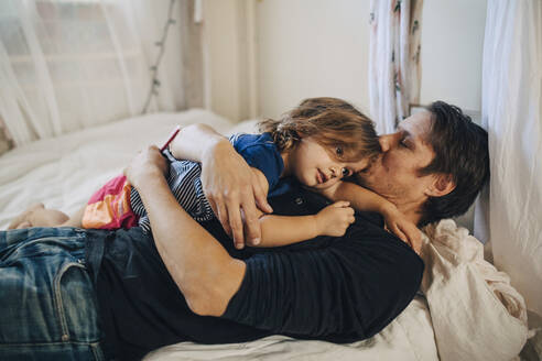 Reifer Vater mit geschlossenen Augen küsst Tochter auf dem Bett liegend - MASF17245