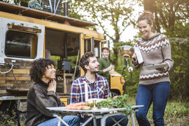 Lächelnde Frau serviert Essen für Freunde, die am Tisch vor einem Wohnwagen sitzen, beim Camping im Wald - MASF17267