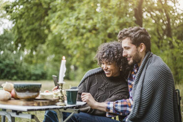 Cheerful young couple sharing mobile phone while wrapped in blanket together during camping in forest - MASF17270