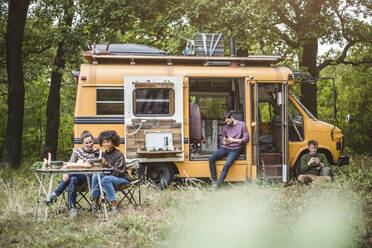 Lächelnde Freundinnen, die am Tisch sitzen, während die Männer beim Camping im Wald ihre Smartphones benutzen - MASF17271