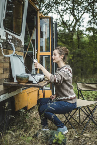 Frau in voller Länge bei der Zubereitung von Kaffee auf dem Herd eines Wohnmobils beim Camping im Wald, lizenzfreies Stockfoto