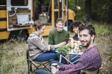 Porträt eines lächelnden Mannes, der mit Freunden am Tisch sitzt, beim Camping im Wald - MASF17284