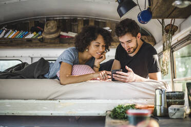 Junges Paar teilt sich ein Smartphone, während es in einem Wohnwagen auf dem Bett liegt - MASF17304