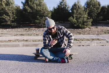 Moderner junger Mann, der auf einem Longboard sitzt und auf sein Mobiltelefon schaut - CAVF76682