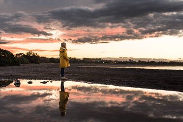 Mädchen in gelbem Mantel steht am Strand bei Sonnenuntergang mit Reflexion - CAVF76757