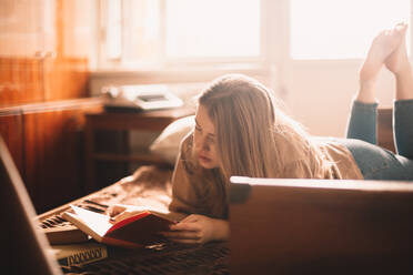 Junge Frau liest ein Buch, während sie zu Hause auf dem Bett liegt - CAVF76807