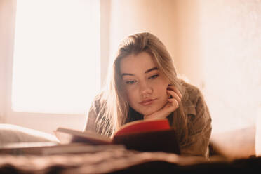 Junge Frau liest ein Buch, während sie zu Hause auf dem Bett liegt - CAVF76808