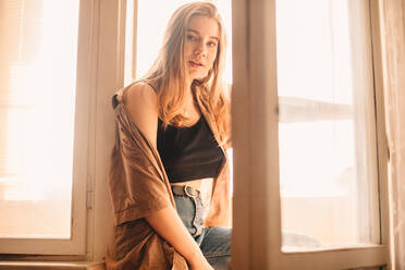 Young woman standing at balcony doorway at home in summer - CAVF76813