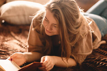 Junge glückliche Frau liest ein Buch, während sie zu Hause auf dem Bett liegt - CAVF76815