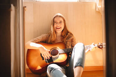 Cheerful young woman holding guitar while sitting on balcony floor - CAVF76817