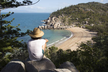 Mann mit lockigem Haar, Hut und Badeanzug, auf einem Felsen, die Bucht bewundernd - CAVF76891