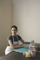 Nachdenkliche Frau in Schürze sitzt mit Behältern und Zutaten für die Zubereitung von Speisen zu Hause - CAVF76895