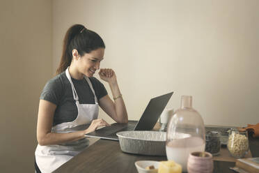 Lächelnde Frau, die am Laptop ein Rezept liest, während sie am Tisch Essen zubereitet - CAVF76899