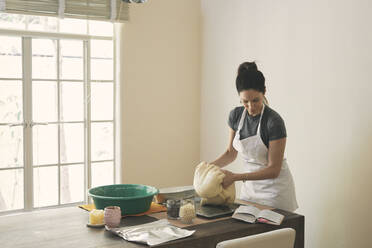 Woman kneading dough while reading recipe in book at table - CAVF76901