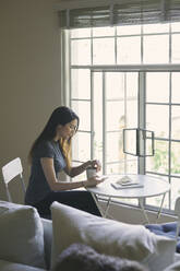 Woman using mobile phone while having coffee at table in living room - CAVF76912