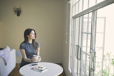 Thoughtful young woman having coffee while looking through window in living room at home - CAVF76914