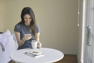 Glückliche Frau, die ein Mobiltelefon benutzt, während sie am Tisch an der Wand im Wohnzimmer Kaffee trinkt - CAVF76916