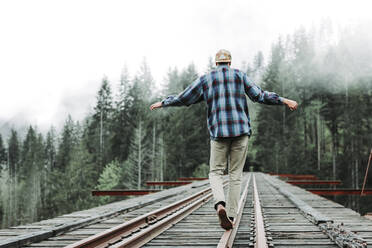 Young man balancing on railroad tracks over bridge in foggy forest - CAVF76966